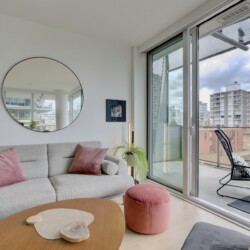 Bright living room with a gray sectional, pink cushions, round wall mirror, and glass balcony doors overlooking city buildings.