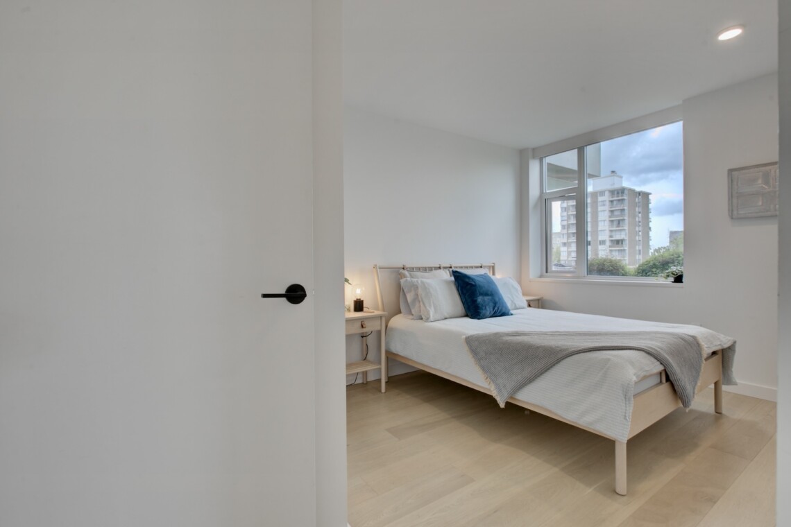 Bright, minimalist bedroom with a wooden bed, white bedding, blue pillow, and a large window revealing city buildings outside.