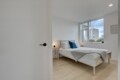 Bright, minimalist bedroom with a wooden bed, white bedding, blue pillow, and a large window revealing city buildings outside.