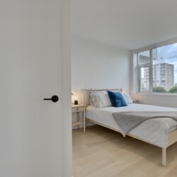 Bright, minimalist bedroom with a wooden bed, white bedding, blue pillow, and a large window revealing city buildings outside.