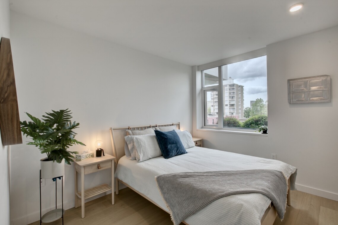 Bright bedroom with a bed, throw pillows, a bedside table lamp, and a large window showing city buildings outside—soft natural light.