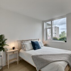 Bright bedroom with a bed, throw pillows, a bedside table lamp, and a large window showing city buildings outside—soft natural light.