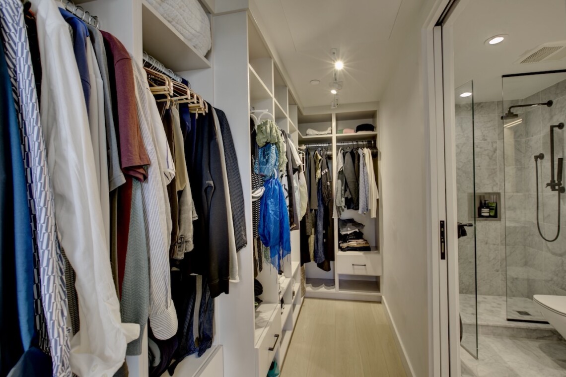 Walk-in closet with organized racks of shirts, jackets, and shelves, adjacent to a glass-enclosed marble bathroom shower.