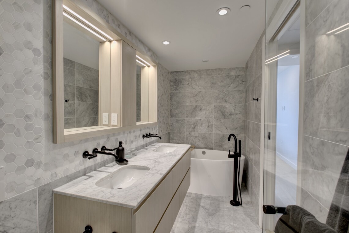 Modern bathroom with a marble double vanity, hex tile backsplash, and a glass shower door in a bright space.