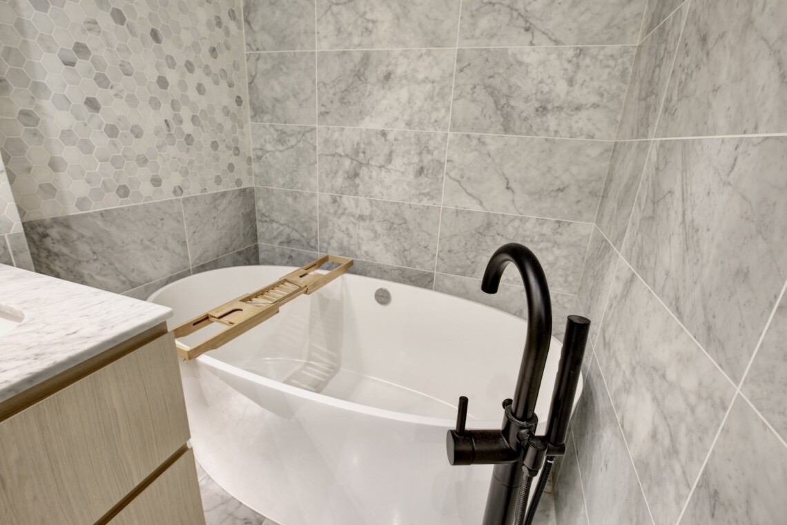 Bathtub with a wooden bath rack inside a marble-tiled bathroom, curved black faucet visible to the right.