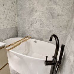 Bathtub with a wooden bath rack inside a marble-tiled bathroom, curved black faucet visible to the right.
