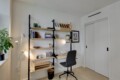 Cozy home office with a tall shelving unit, light wood shelves, books, boxes, and a gray wheeled office chair beside a white desk lamp and plant on the left.