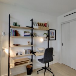 Cozy home office with a tall shelving unit, light wood shelves, books, boxes, and a gray wheeled office chair beside a white desk lamp and plant on the left.