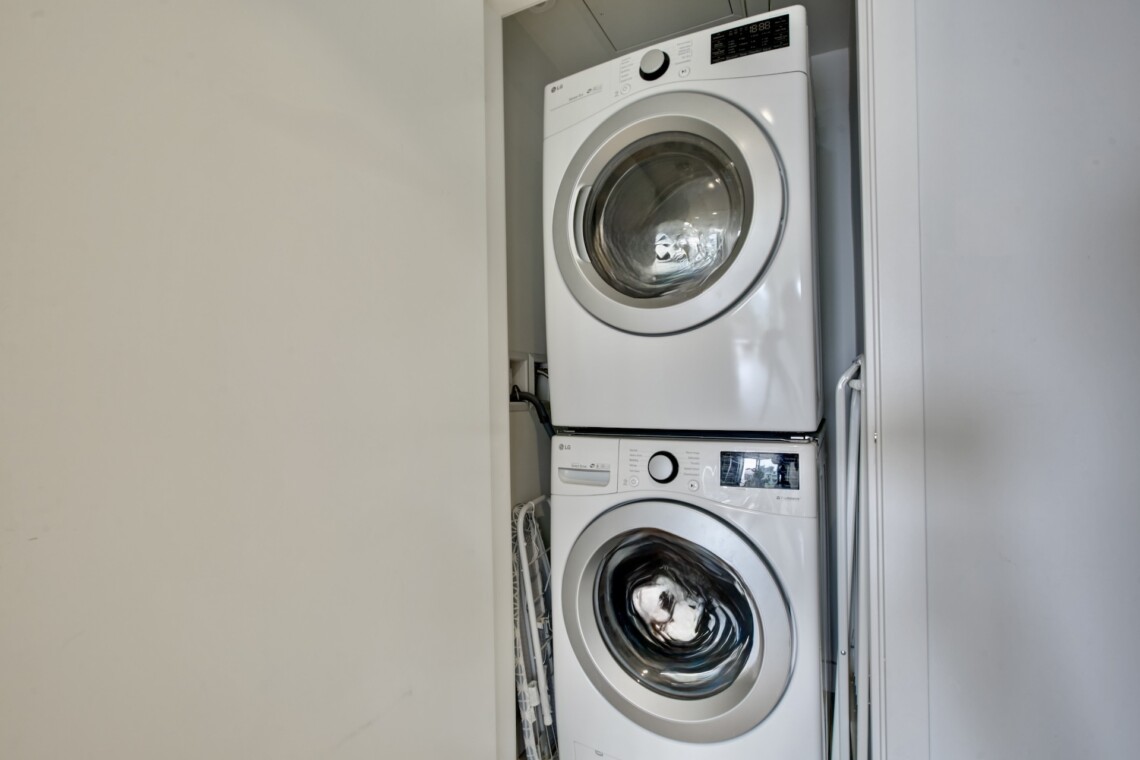 Stacked white front-loading washer and dryer in a narrow laundry closet with doors open to the side.