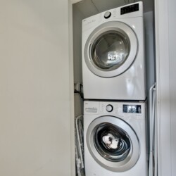 Stacked white front-loading washer and dryer in a narrow laundry closet with doors open to the side.