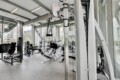 Modern gym interior with silver weight machines and cable stations, large windows overlooking city buildings in the background.