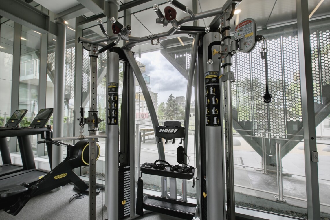 Modern gym with a multi-station weight machine near large glass walls; treadmill visible on the left and various cables and pulleys in view.
