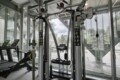 Modern gym with a multi-station weight machine near large glass walls; treadmill visible on the left and various cables and pulleys in view.