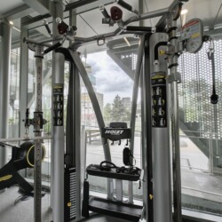 Modern gym with a multi-station weight machine near large glass walls; treadmill visible on the left and various cables and pulleys in view.