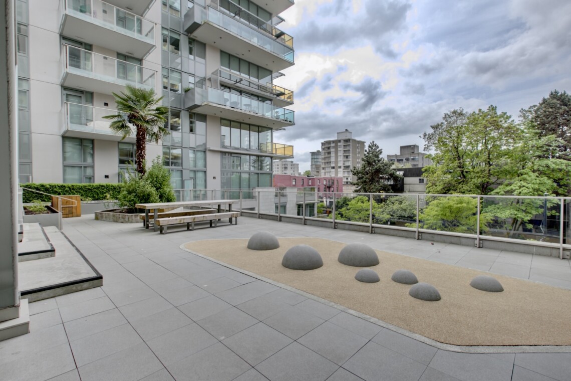 Rooftop courtyard of a modern apartment building with glass balconies, benches, a palm tree, and a sandy play area with dome-shaped sculptures.