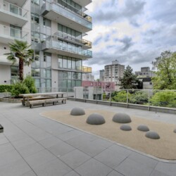 Rooftop courtyard of a modern apartment building with glass balconies, benches, a palm tree, and a sandy play area with dome-shaped sculptures.