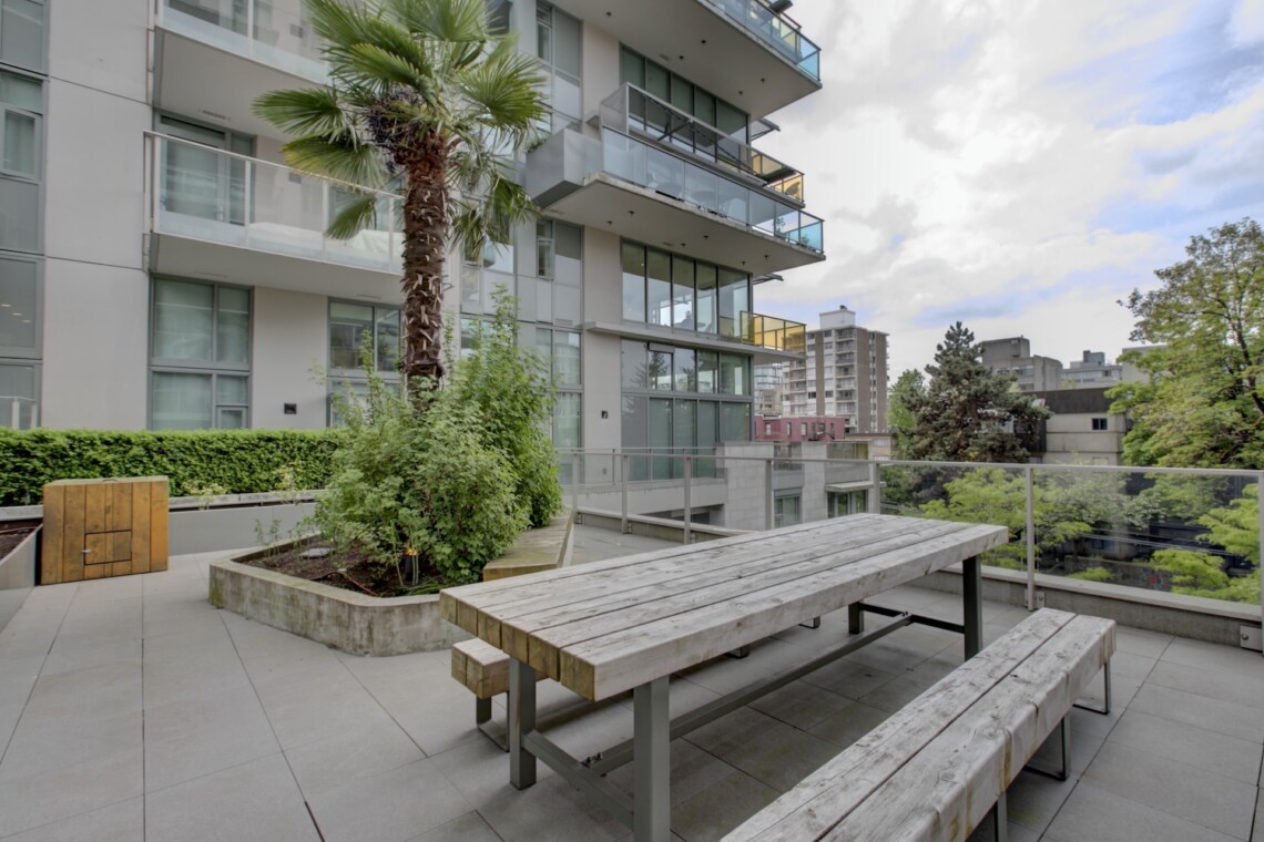 Outdoor terrace with long wooden benches and a tall palm tree, adjacent to a modern apartment building with glass balconies and greenery in planters.