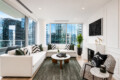 Bright modern living room with a white sectional, green and black-and-white cushions, and floor-to-ceiling city-view windows.
