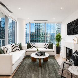 Bright modern living room with a white sectional, green and black-and-white cushions, and floor-to-ceiling city-view windows.