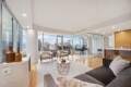 Modern living room with a gray sofa, beige chairs, and a round wire coffee table, overlooking a city skyline through large windows.