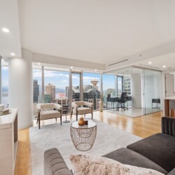Modern living room with a gray sofa, beige chairs, and a round wire coffee table, overlooking a city skyline through large windows.