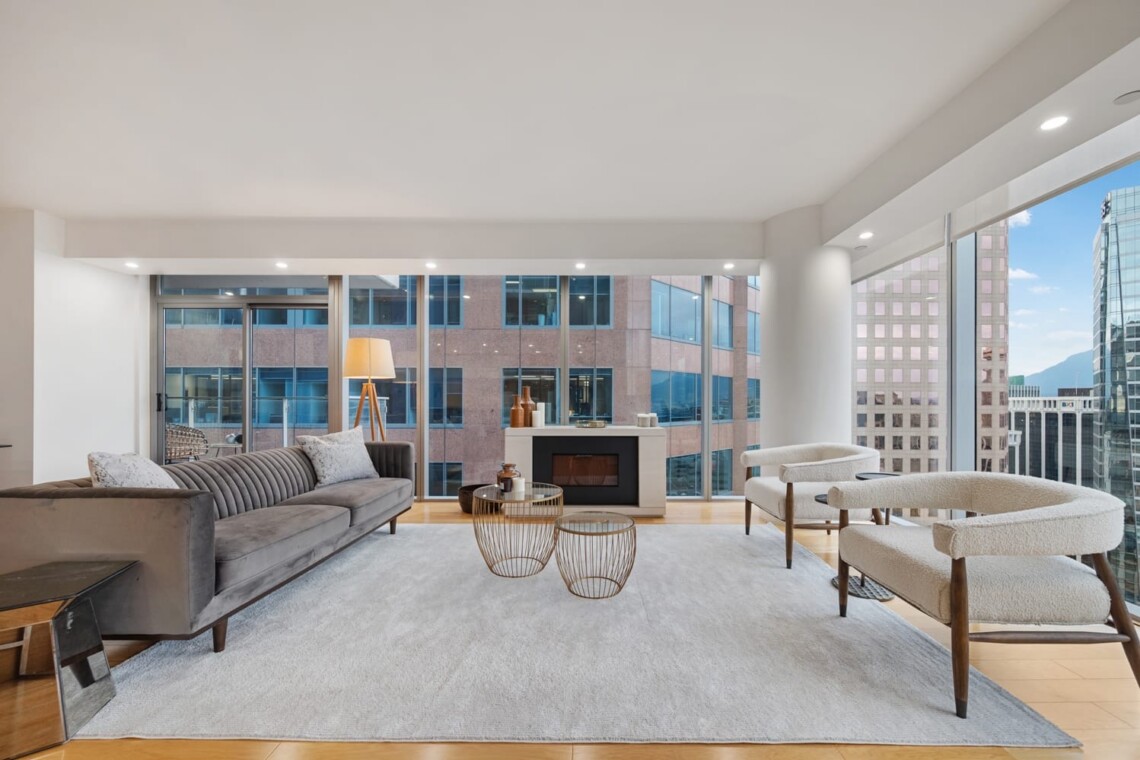 Modern living room with a grey sofa, two white boucle chairs, and a fireplace overlooking city skyscrapers through large floor-to-ceiling windows in a bright, airy space.