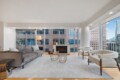 Modern living room with a grey sofa, two white boucle chairs, and a fireplace overlooking city skyscrapers through large floor-to-ceiling windows in a bright, airy space.
