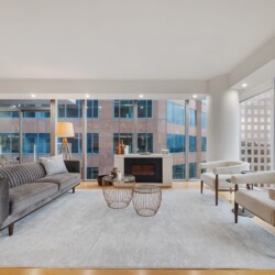 Modern living room with a grey sofa, two white boucle chairs, and a fireplace overlooking city skyscrapers through large floor-to-ceiling windows in a bright, airy space.
