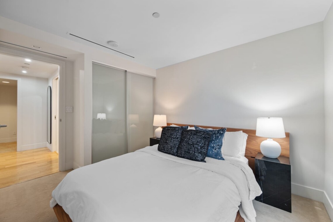 Bedroom with a bed, white linens, and blue decorative pillows, flanked by two lamps on dark side tables and a frosted glass wardrobe behind.