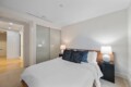 Bedroom with a bed, white linens, and blue decorative pillows, flanked by two lamps on dark side tables and a frosted glass wardrobe behind.