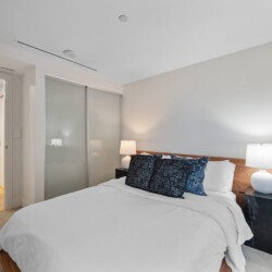 Bedroom with a bed, white linens, and blue decorative pillows, flanked by two lamps on dark side tables and a frosted glass wardrobe behind.