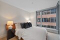 Modern bedroom with a wooden bed, white bedding, and dark throw pillows, flanked by two lamps near a large window.