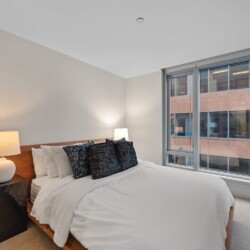 Modern bedroom with a wooden bed, white bedding, and dark throw pillows, flanked by two lamps near a large window.