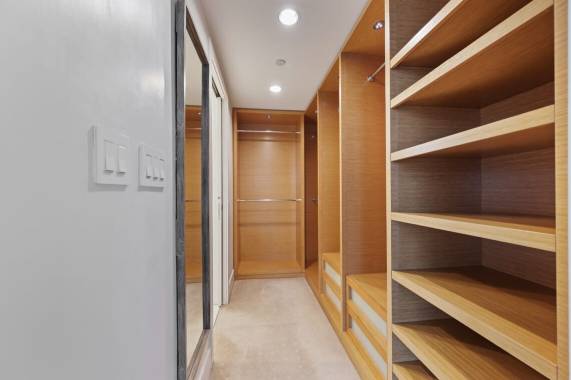 Narrow walk-in closet with built-in wooden shelves and hanging rods, illuminated by ceiling lights