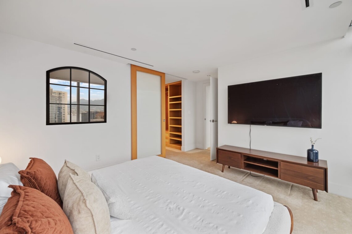 Bright modern bedroom with a wall-mounted TV above a wooden console, a white bed with rust and beige pillows, and an open wooden shelving closet next to a frosted-glass sliding door; a cityscape window on the left.