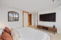 Bright modern bedroom with a wall-mounted TV above a wooden console, a white bed with rust and beige pillows, and an open wooden shelving closet next to a frosted-glass sliding door; a cityscape window on the left.