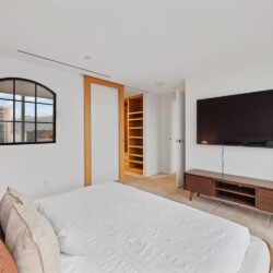 Bright modern bedroom with a wall-mounted TV above a wooden console, a white bed with rust and beige pillows, and an open wooden shelving closet next to a frosted-glass sliding door; a cityscape window on the left.