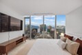 Bedroom with a large window overlooking a city skyline, king-size bed with white linens and beige pillows beside a wooden console and TV massing the wall.