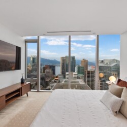 Bedroom with a large window overlooking a city skyline, king-size bed with white linens and beige pillows beside a wooden console and TV massing the wall.