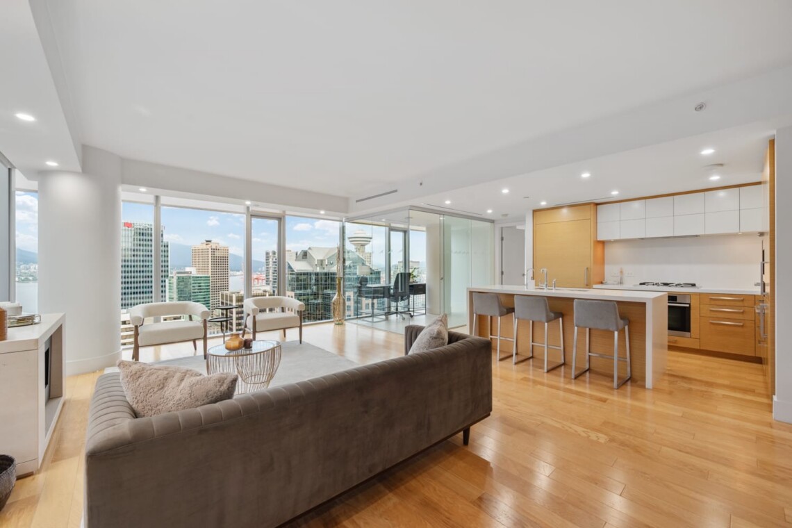 Open-concept living area with a gray sofa, light wood floors, and a modern kitchen, all overlooking a city skyline through floor-to-ceiling windows and a balcony.