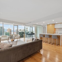 Open-concept living area with a gray sofa, light wood floors, and a modern kitchen, all overlooking a city skyline through floor-to-ceiling windows and a balcony.