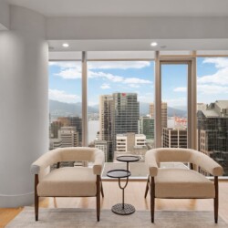 Modern high-rise living area with two beige curved chairs facing large windows that reveal a city skyline and harbor view.