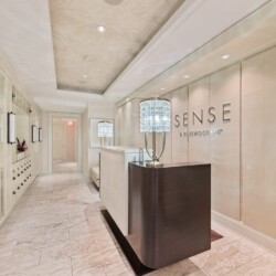 Reception area of Sense Rosewood Spa with a dark curved front desk and crystal lamp centerpiece, marble floors, and glass display cabinets on the left.