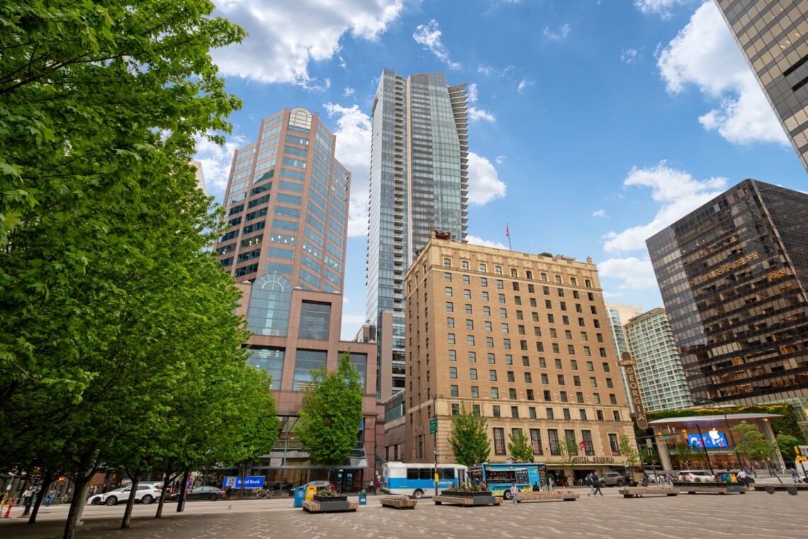 City square with tall modern buildings, green trees on the left, and a bright blue sky behind them.