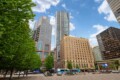 City square with tall modern buildings, green trees on the left, and a bright blue sky behind them.