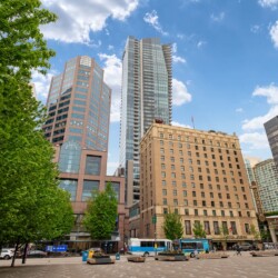 City square with tall modern buildings, green trees on the left, and a bright blue sky behind them.