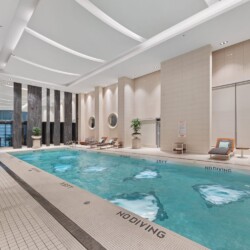 Indoor hotel pool area with lounge chairs along the water and tall dark stone columns under a bright ceiling.