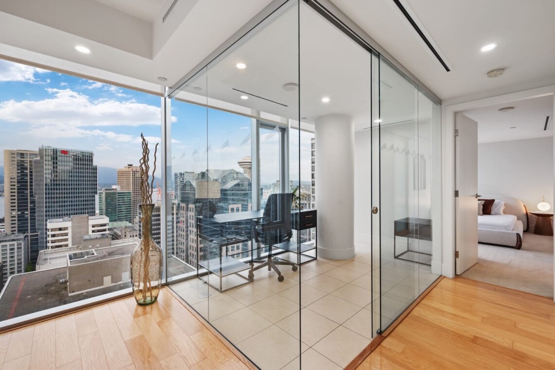 Modern glass-walled office with a desk and chair, overlooking a city skyline from a high floor, next to a bedroom.