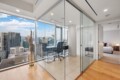 Modern glass-walled office with a desk and chair, overlooking a city skyline from a high floor, next to a bedroom.