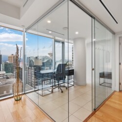 Modern glass-walled office with a desk and chair, overlooking a city skyline from a high floor, next to a bedroom.